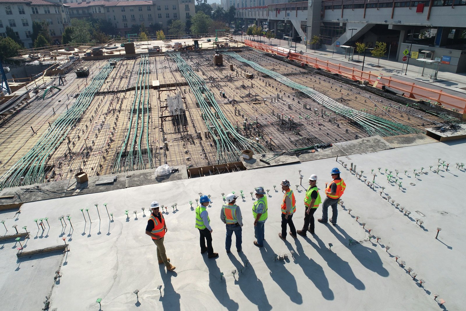 A superintendent and crew in hard hats and high-visibility vests walking a freshly poured deck beside a post-tension slab and rebar mat.